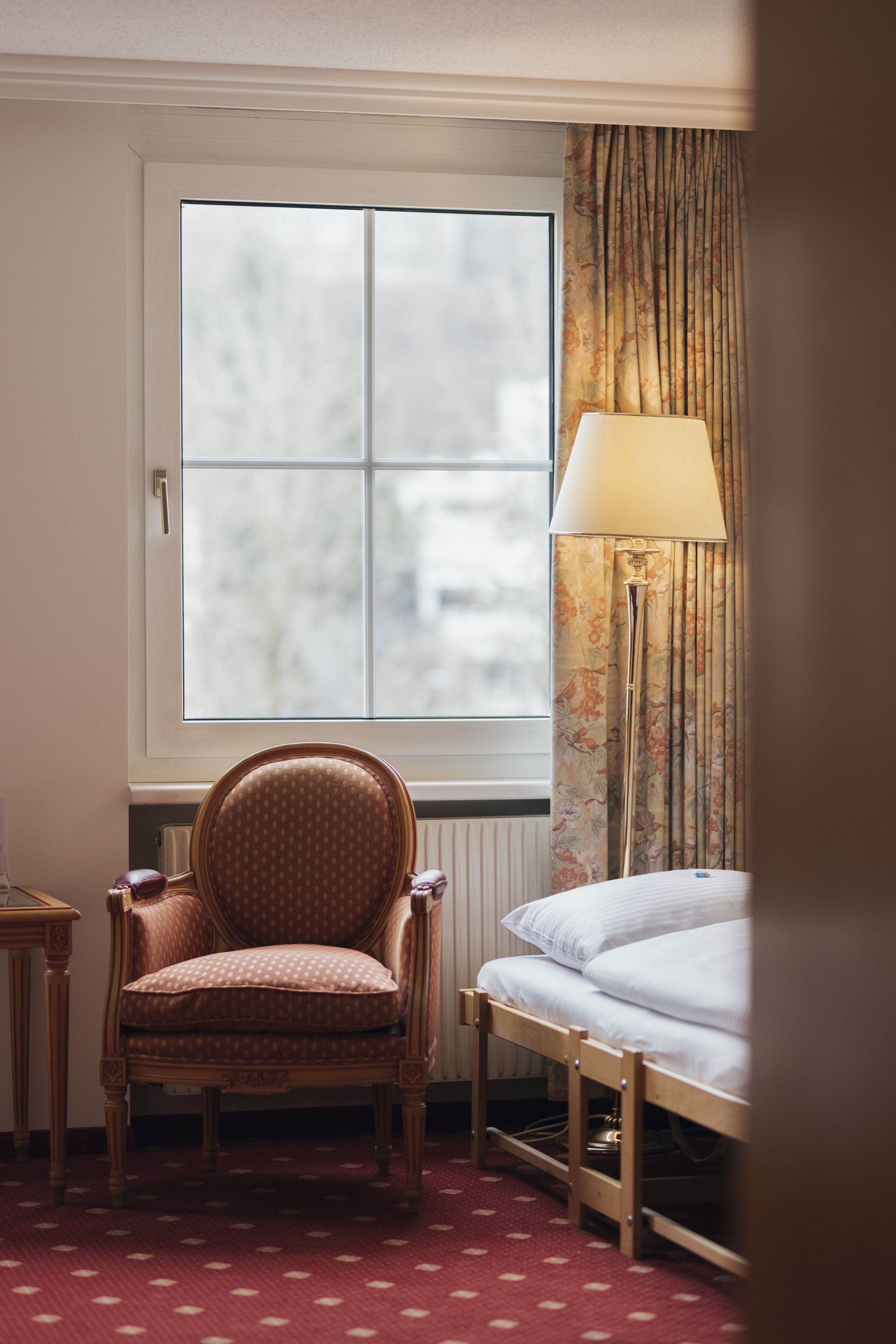 A hotel room with a large window, a chair, a bedside table, a lamp, and a bed.