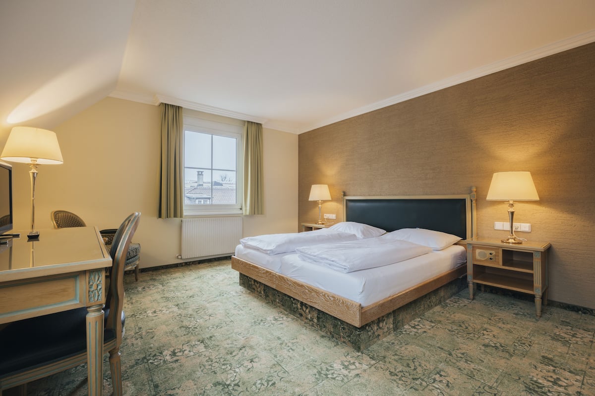 Une chambre d’hôtel comprenant un lit, deux tables de chevet avec des lampes, un bureau avec une chaise et une fenêtre avec des rideaux.