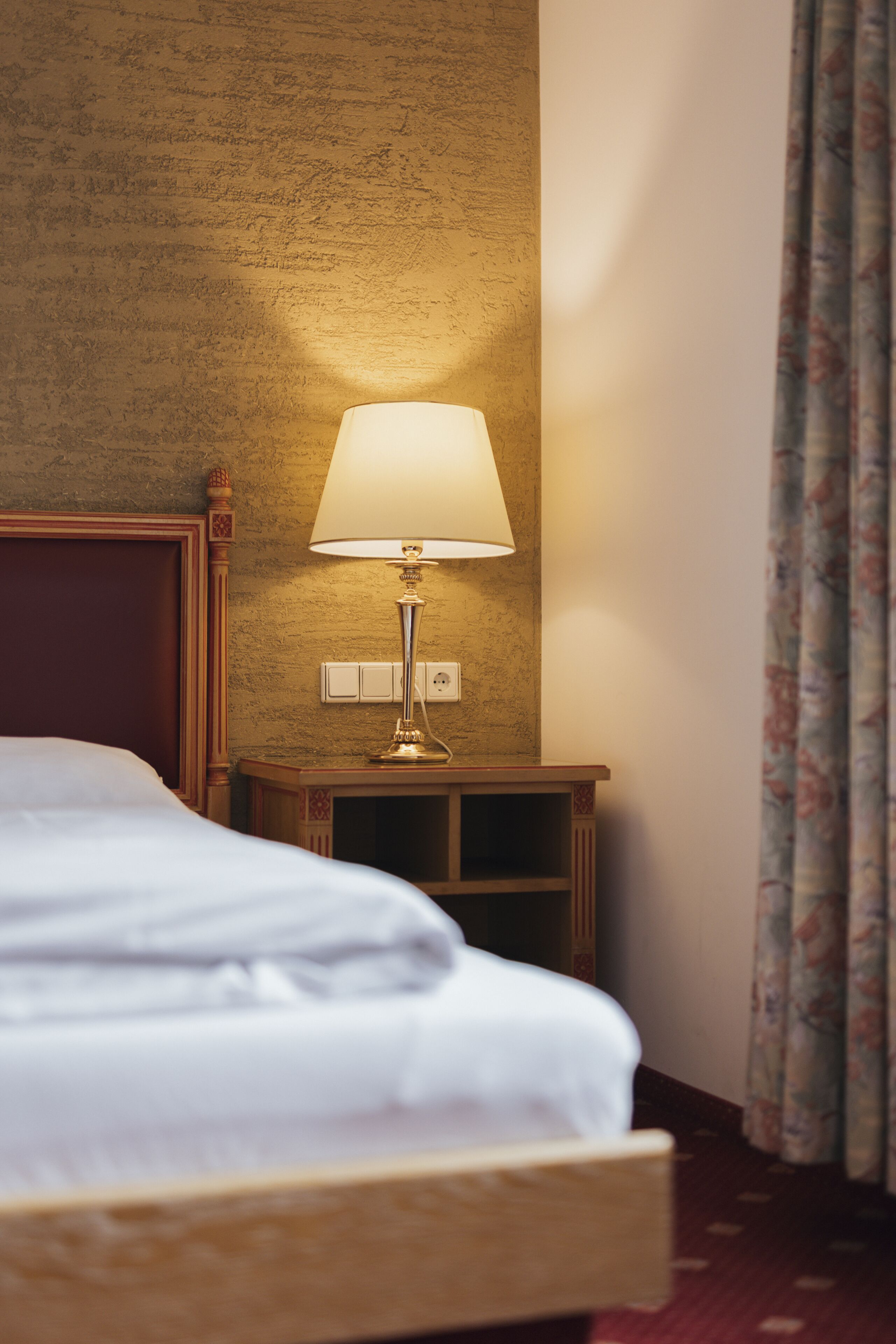 A hotel room with a bed, a nightstand with a lamp, and a power outlet on the wall.
