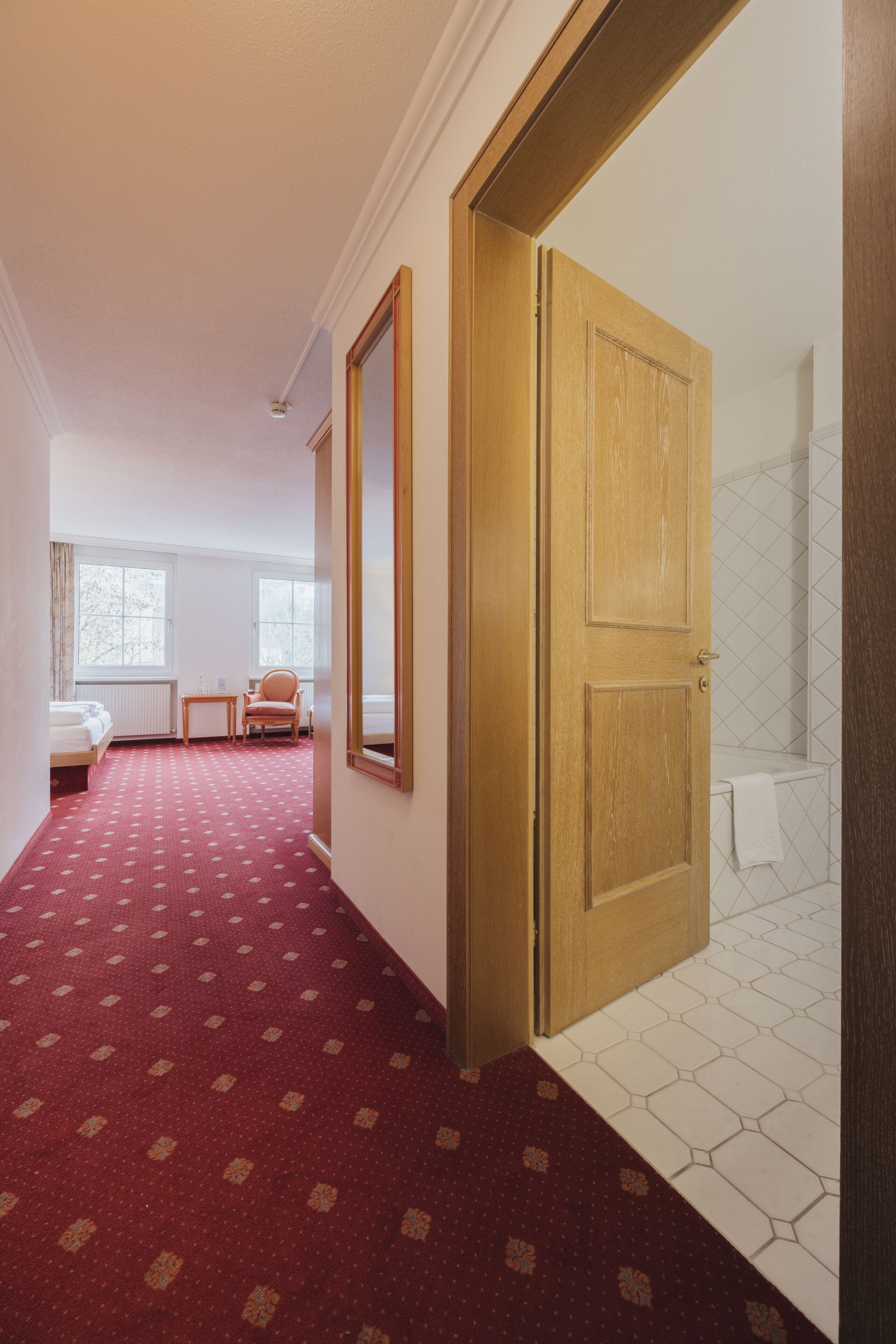 A hotel room with a red carpet, a wooden door, a white radiator, a bed, a chair, and a window.