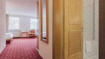 Une chambre d’hôtel avec un tapis rouge, une porte en bois, un radiateur blanc, un lit, une chaise et une fenêtre.