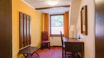A hotel room with a wooden bench, a chair, a table with a lamp, a window with curtains, and a mirror on the wall.