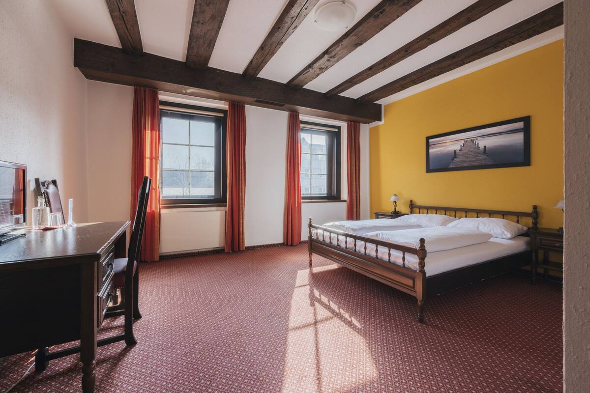 Une chambre avec un lit en bois, un bureau et une chaise. Il y a des fenêtres avec des rideaux, un tableau au mur et un tapis à motifs.