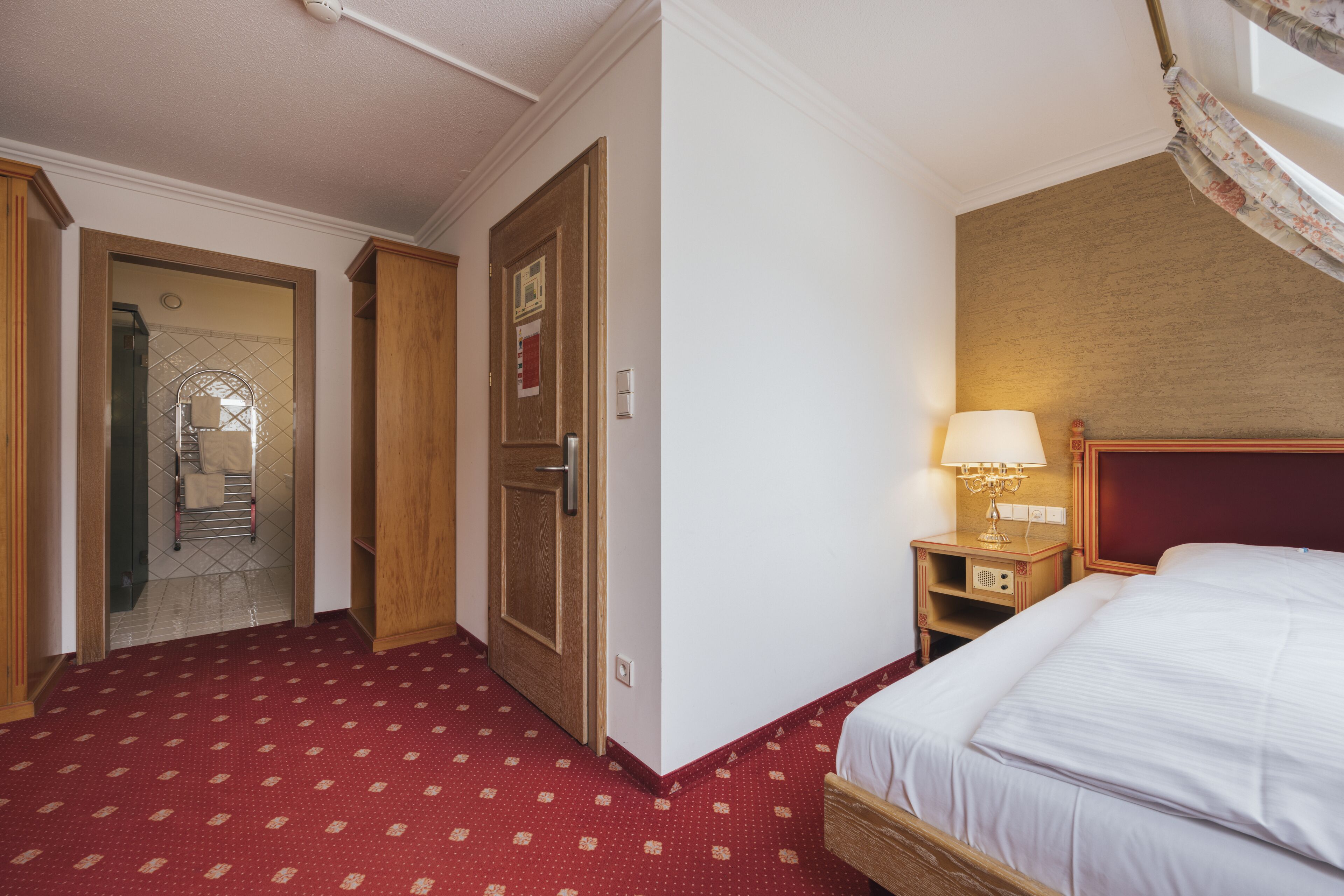Una habitación de hotel con cama, mesita de noche con lámpara, armario de madera y puerta que da a un pasillo.
