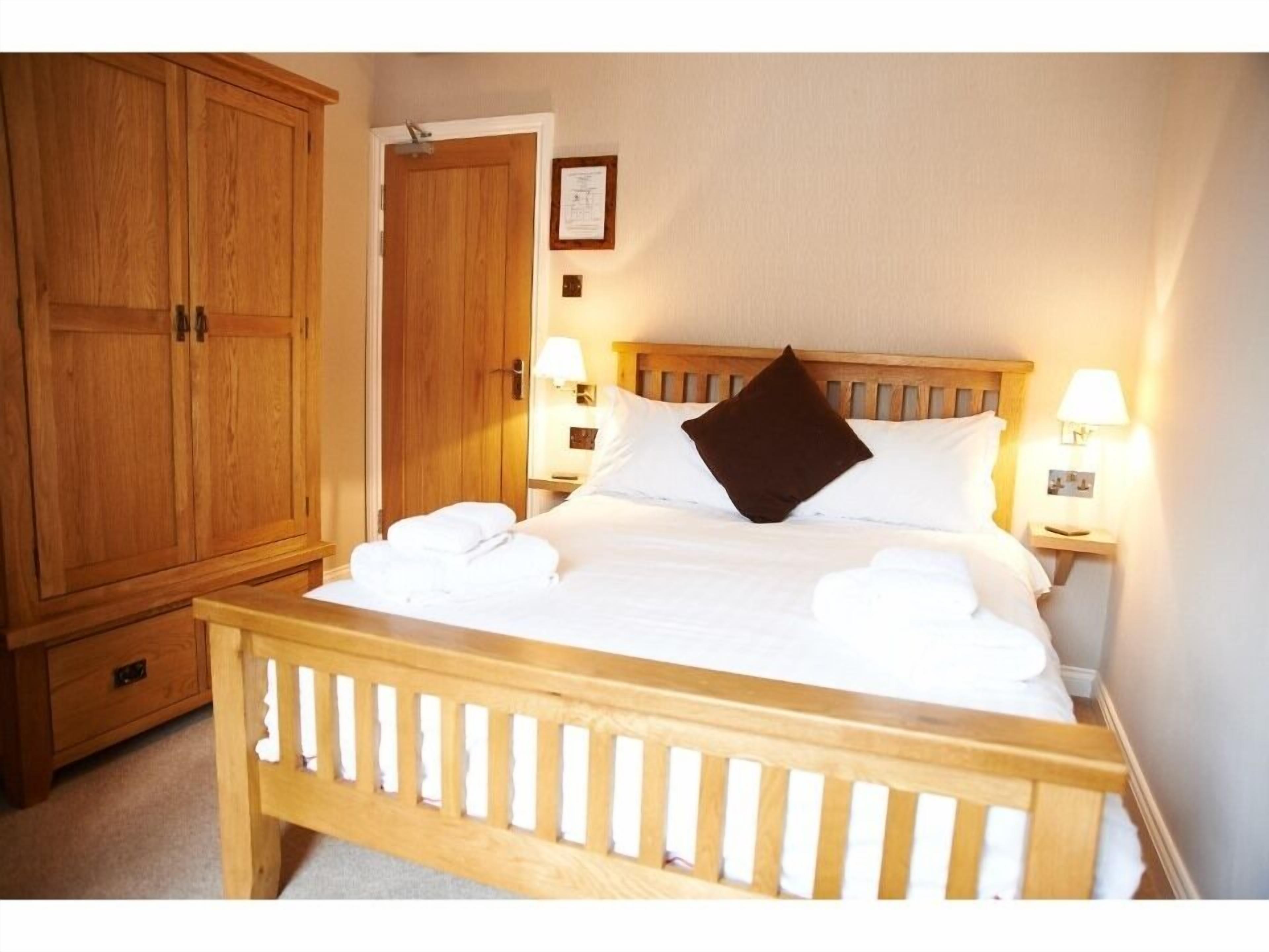 A neatly made bed with white linens and a brown pillow, wooden headboard, two bedside lamps, and a wooden wardrobe.