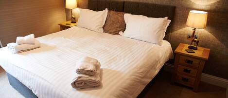 Un lit bien fait, avec du linge de lit blanc et des oreillers, deux tables de chevet avec des lampes, et une serviette pliée posée sur le lit.