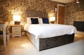 A bedroom with a large bed, two bedside tables with lamps, a wooden nightstand, and a stone-patterned wall.