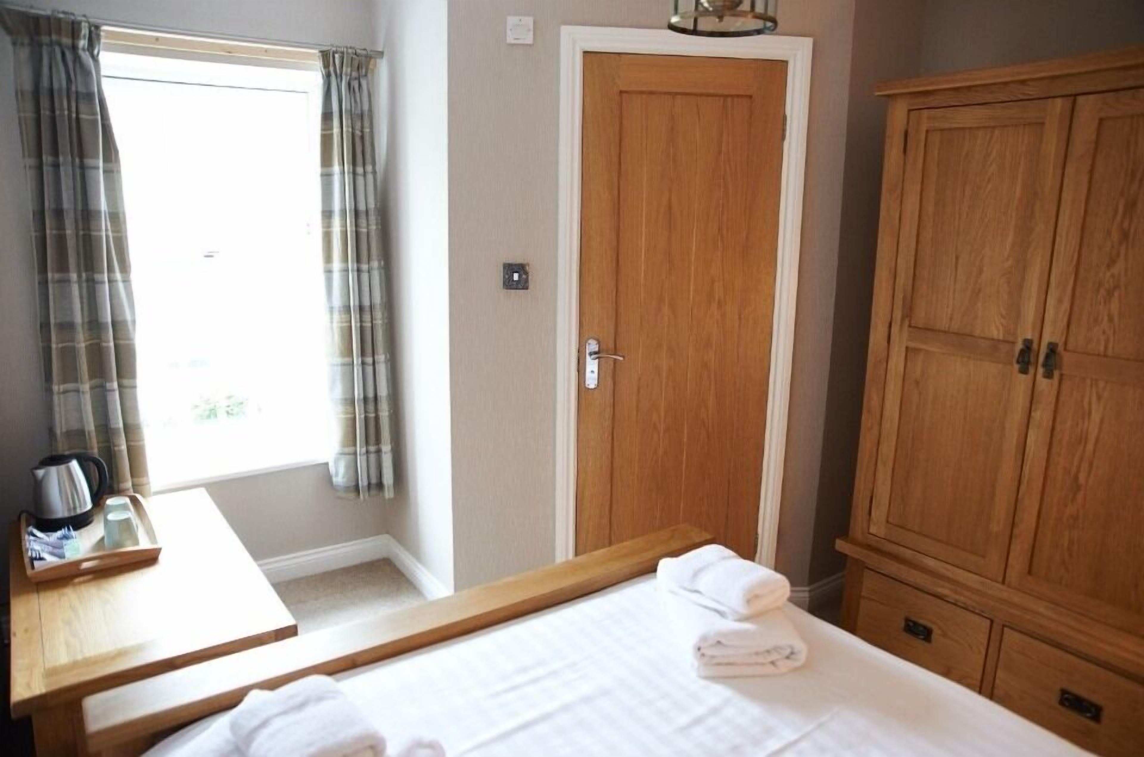 Een hotelkamer met een bed, een houten kledingkast, een nachtkastje met waterkoker en dienblad, en een raam met gordijnen.