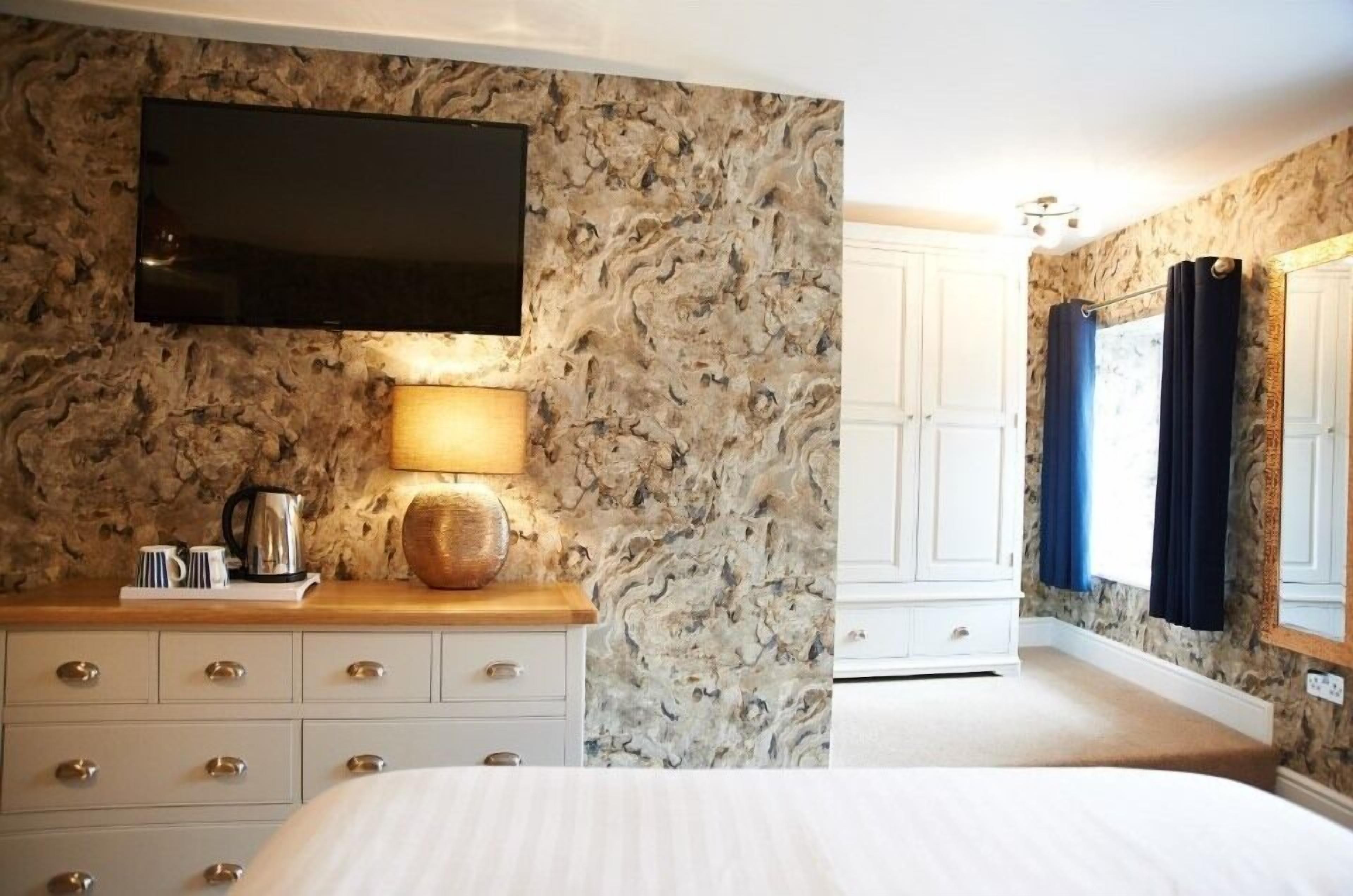 Camera da letto con parete decorata con motivi in marmo, una TV a schermo piatto montata sopra una cassettiera in legno, un letto con biancheria bianca e una finestra con tende blu.