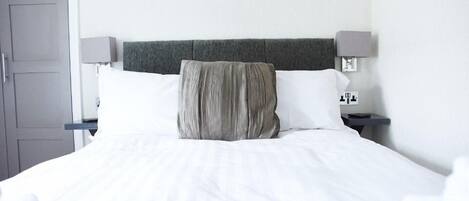 Una cama bien hecha con ropa blanca y un cabecero gris. Hay dos mesitas de noche, una con una lámpara y un pequeño reloj.