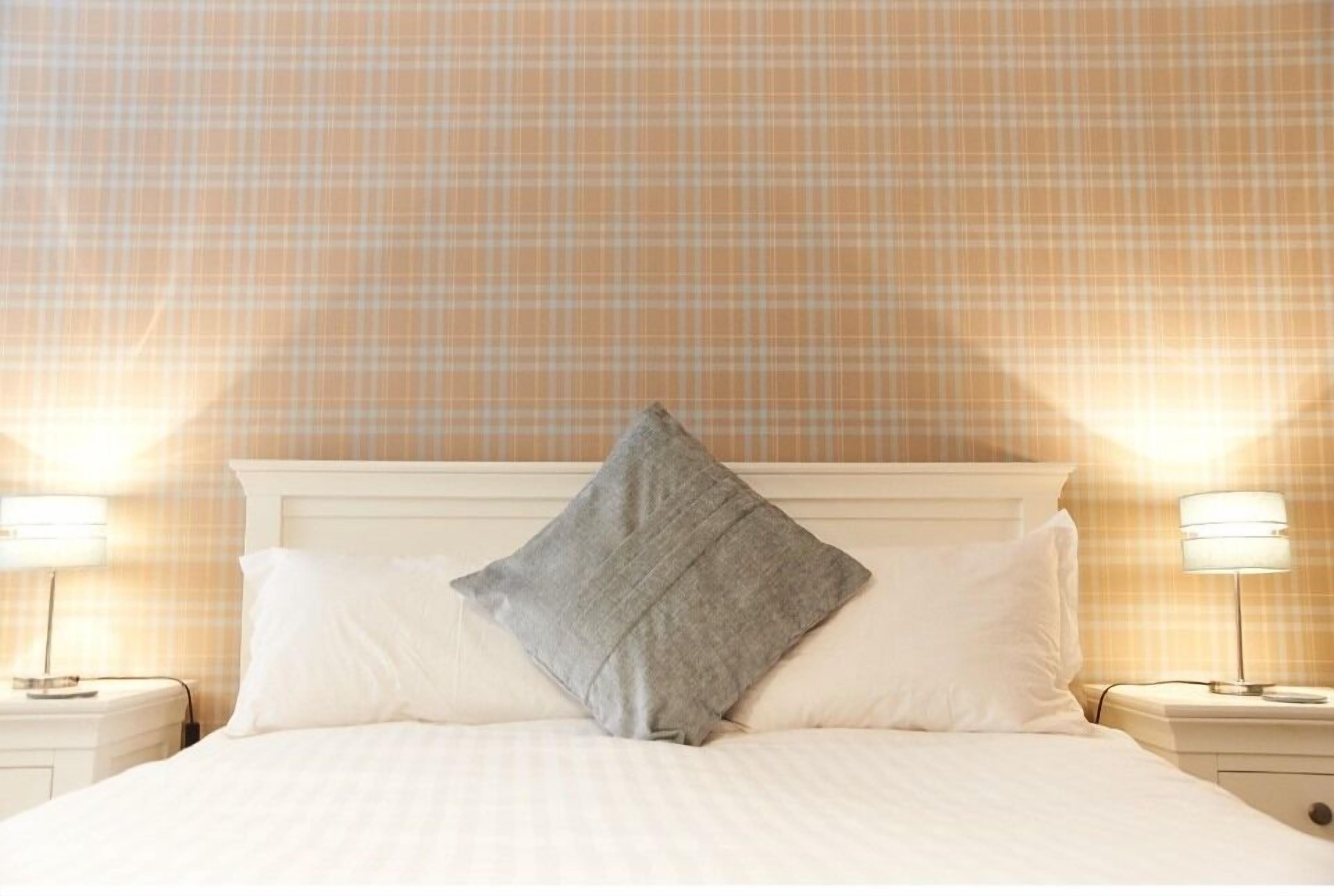 Una cama bien hecha con un almohadón gris y almohadas blancas, flanqueada por dos lámparas de noche sobre mesitas blancas.