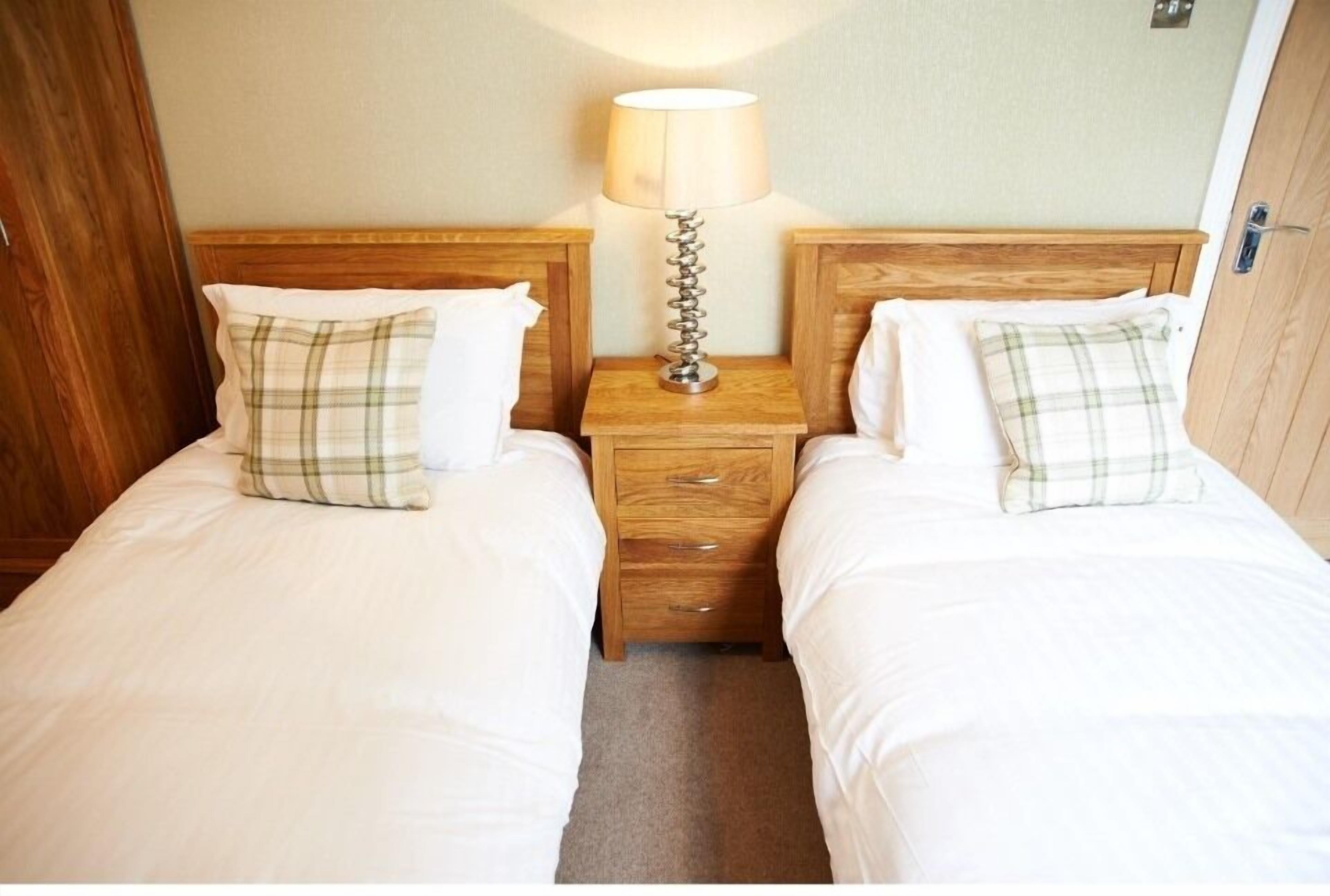 A double room with two beds, each with a wooden headboard and a bedside table. There is a lamp on the nightstand between the beds.