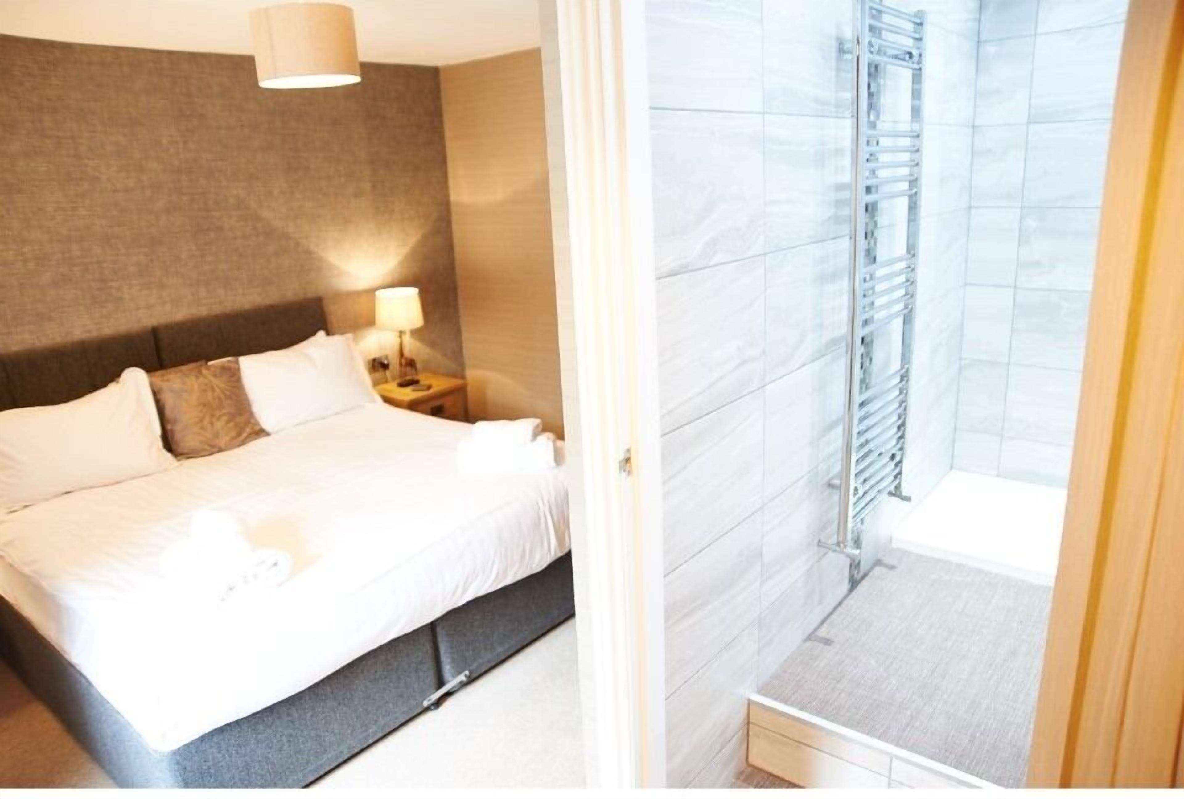 A hotel room with a bed, nightstand, lamp, and a view of a bathroom with a shower and a towel rack.