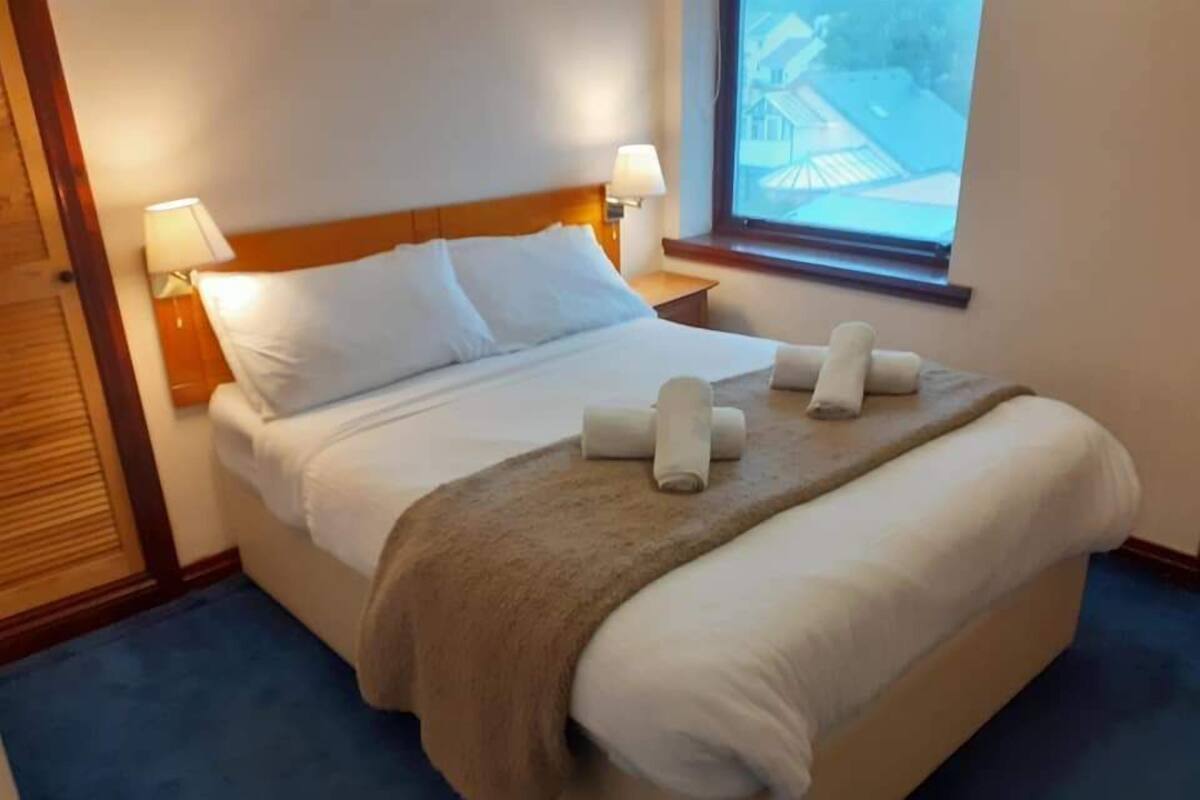 Een netjes opgemaakt bed met witte lakens, een bruine plaid en twee opgerolde handdoeken. Twee bedlampen en een raam met uitzicht op gebouwen.