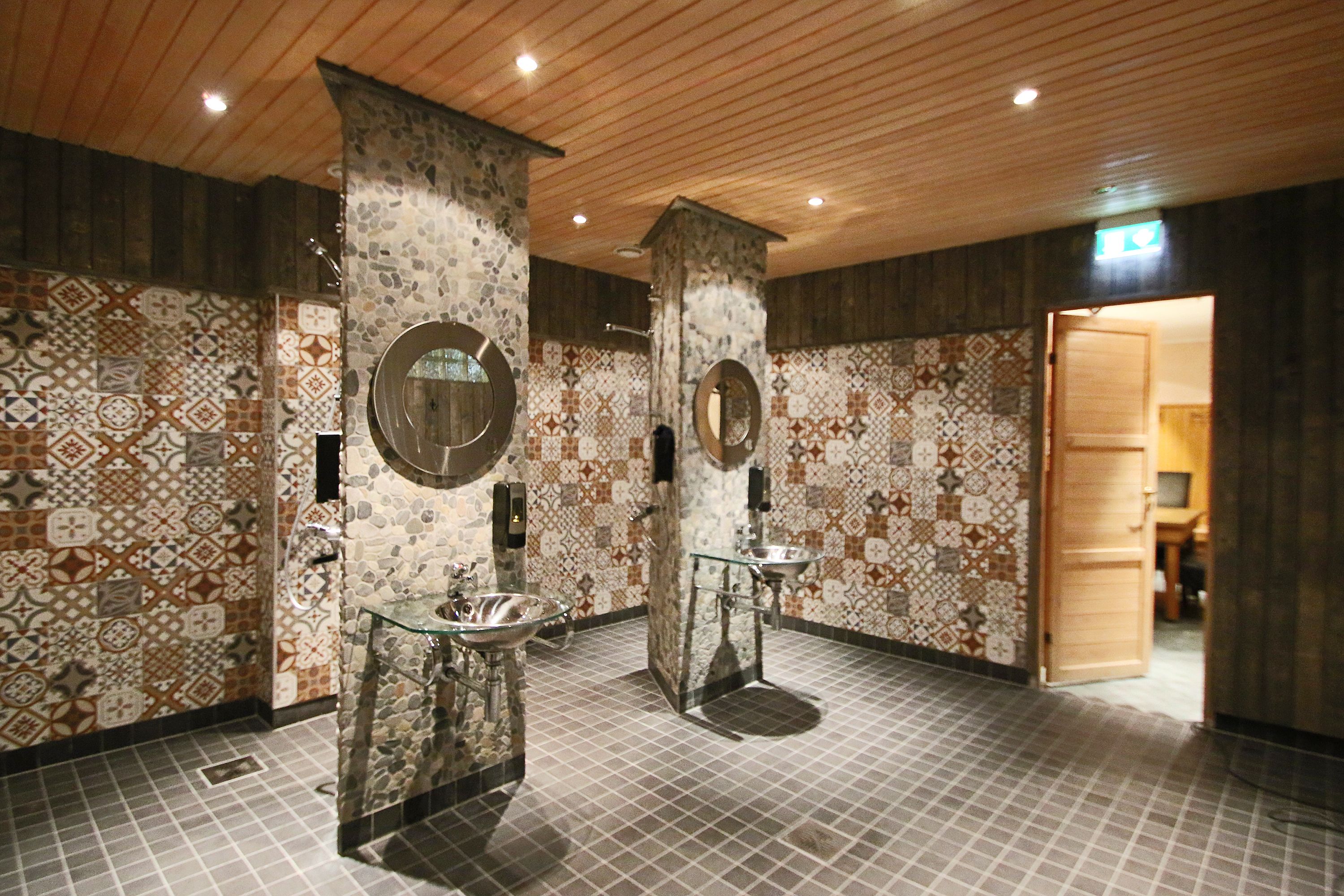 Un baño público con paredes de baldosas estampadas, dos lavamanos con frentes de vidrio y paneles de techo de madera.