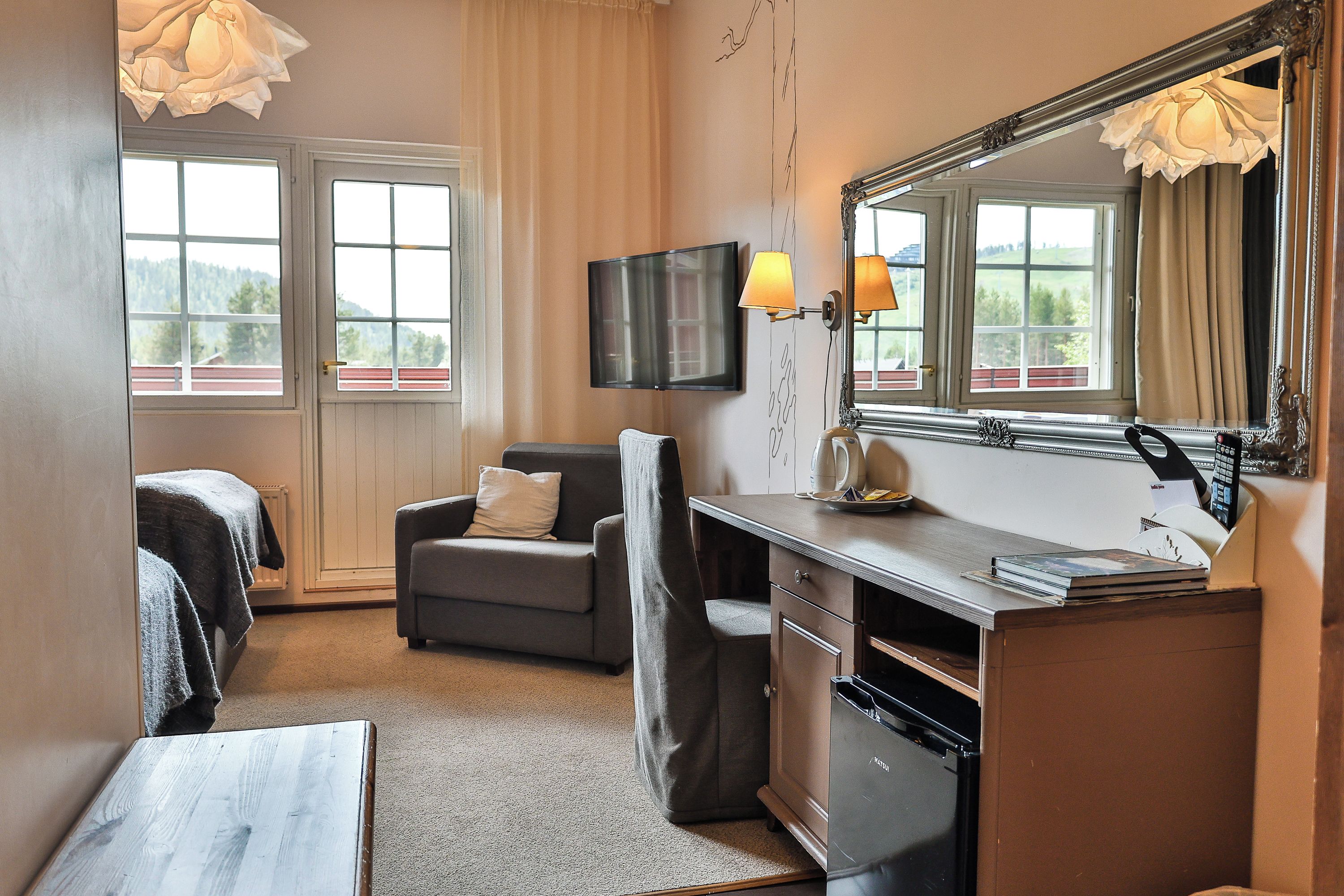 A hotel room with a bed, a desk with a mirror, a television, and a view of the outdoors through the windows.