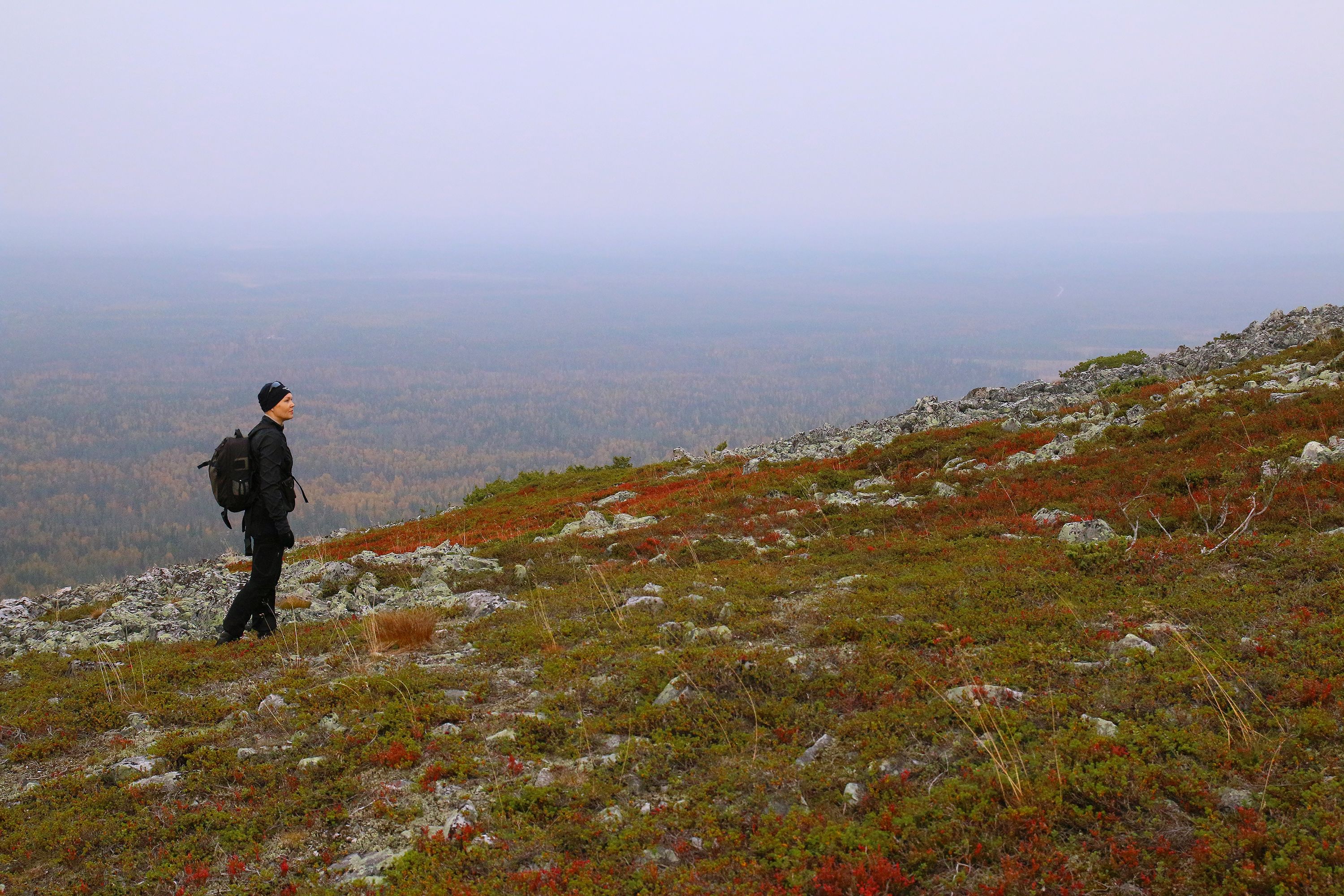 A person standing on a rocky hillside with a backpack, overlooking a forested valley.