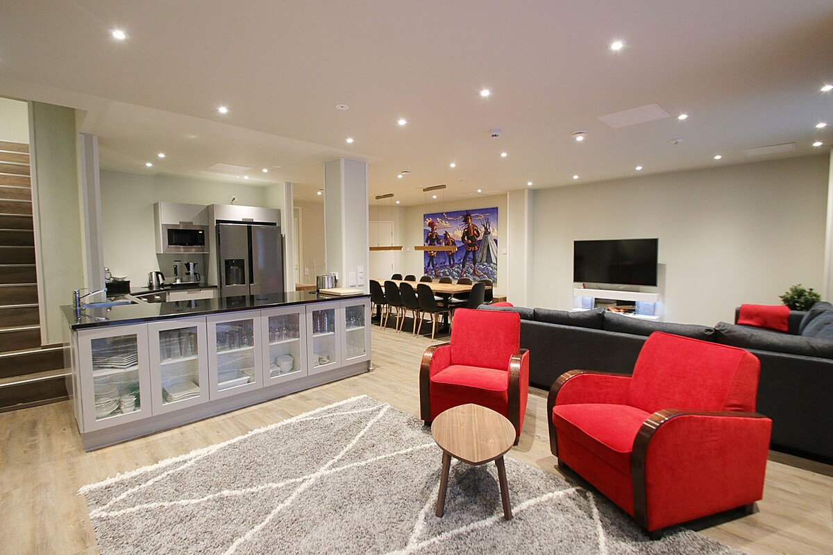 A modern living room with a red sofa, a coffee table, and a TV. There is a kitchen area with a refrigerator and a dining table with chairs.