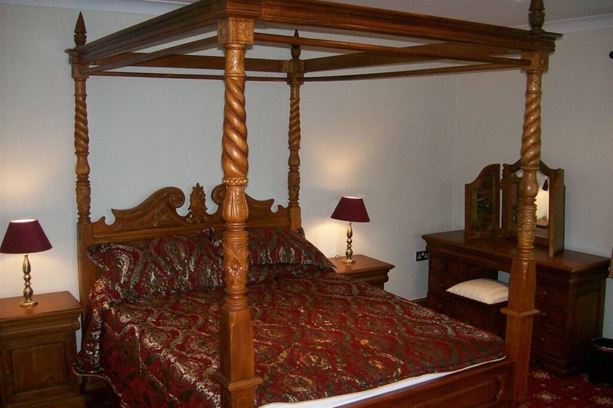 A four-poster bed with a red and gold patterned bedspread, two bedside tables with lamps, and a wooden nightstand with a mirror.