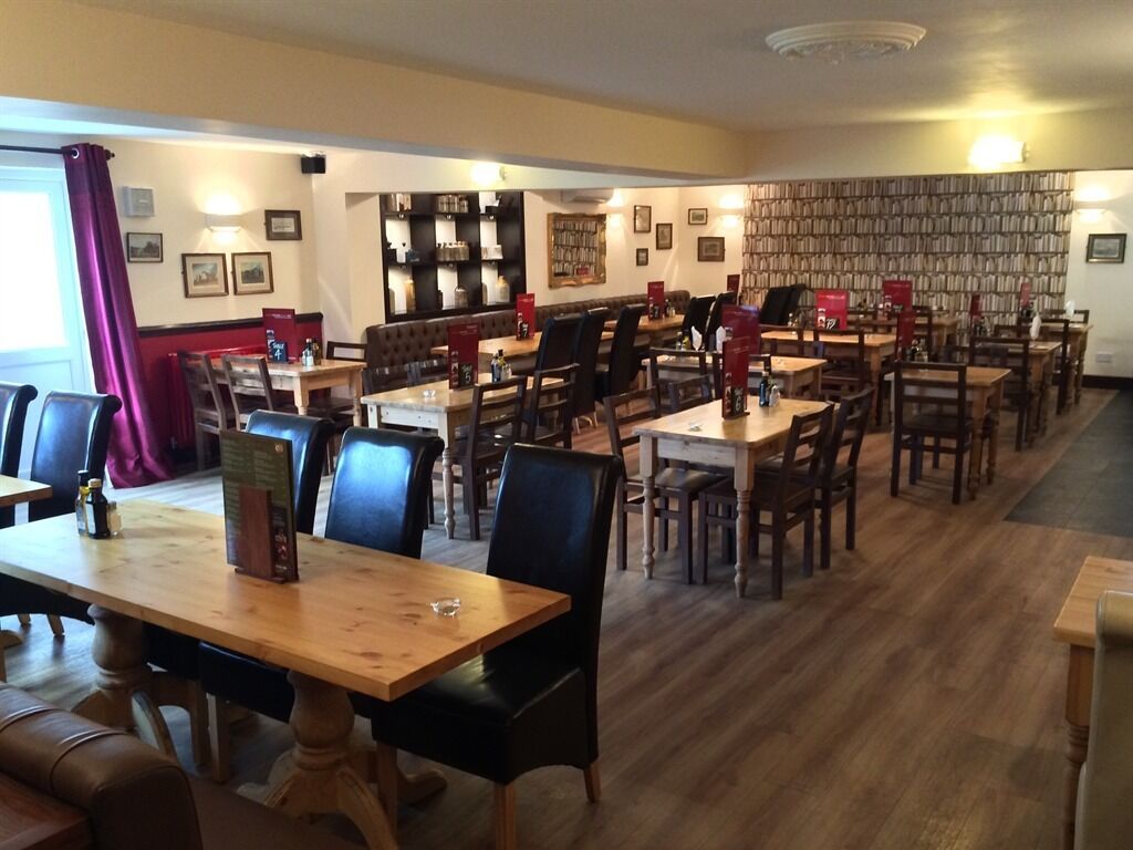 A restaurant with multiple wooden tables and chairs, a bookshelf wall, and framed pictures on the walls.