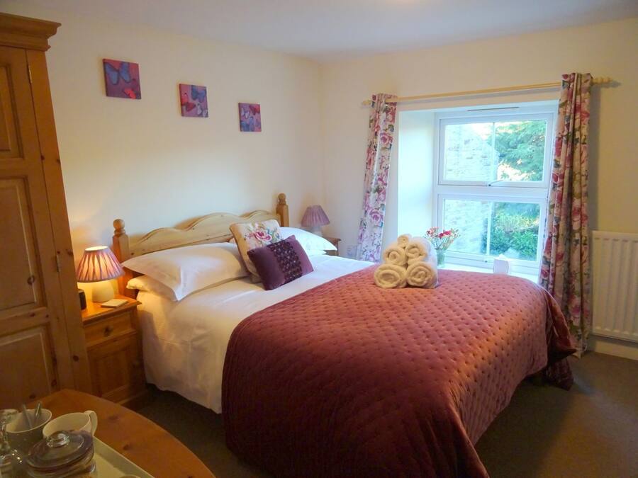 A neatly made bed with a red quilt, a bedside lamp, and a window with floral curtains.