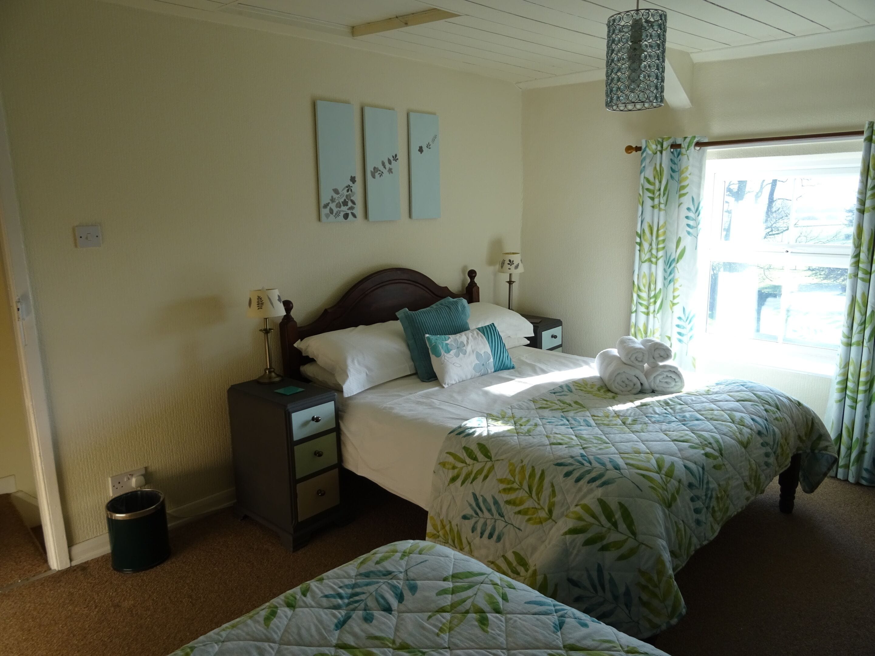 A bedroom with a bed, two bedside tables, a nightstand, a lamp, a window with curtains, and a wall with three framed pictures.