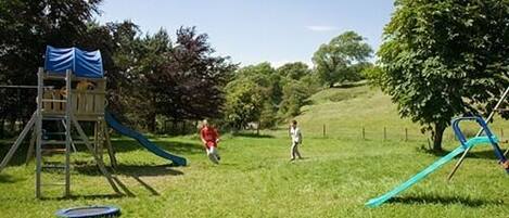 A playground with a slide and climbing structure, surrounded by grass and trees.