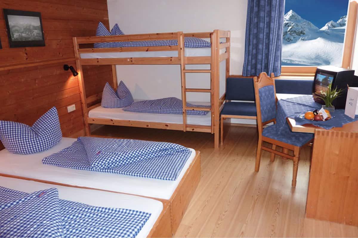 Quarto com beliche, roupa de cama xadrez, escrivaninha com cadeira e janela com vista para as montanhas.