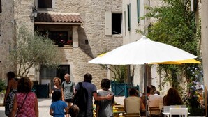 Una estrecha calle adoquinada con edificios de piedra, mesas al aire libre y gente caminando.
