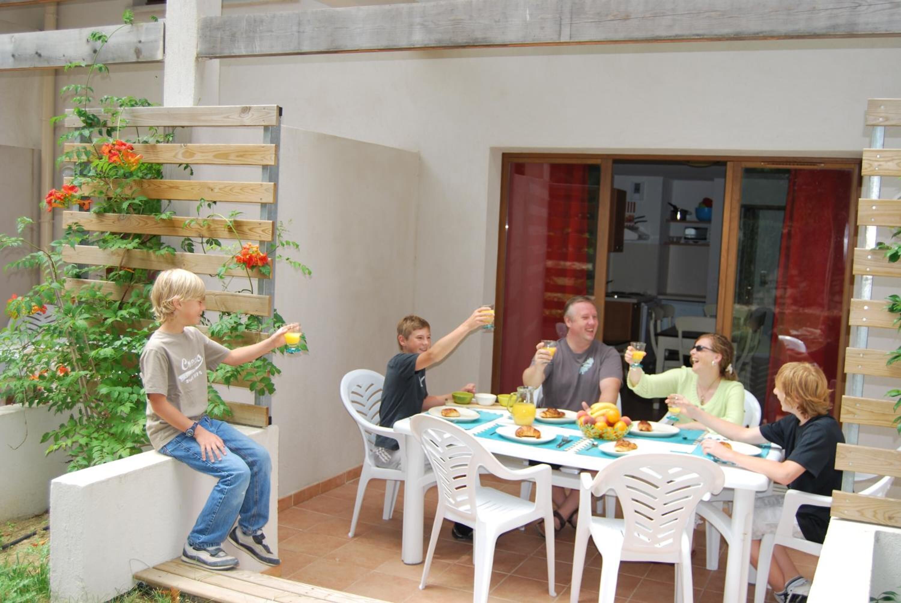 A group of people dining at an outdoor table with white chairs, surrounded by a wooden fence and potted plants.