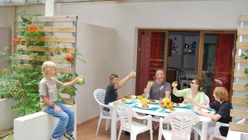 A group of people dining at an outdoor table with white chairs, surrounded by a wooden fence and potted plants.