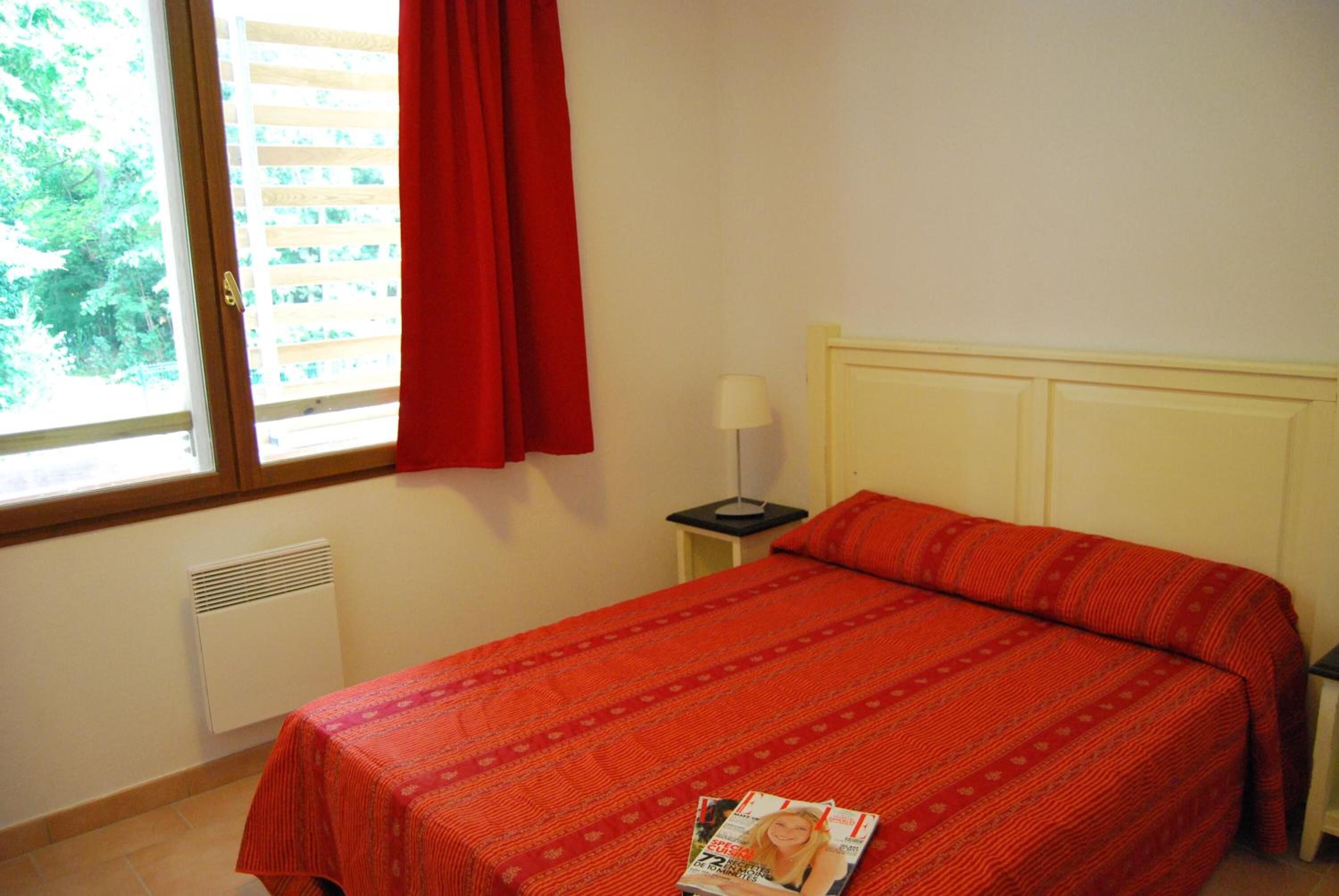 Une chambre avec un lit, une fenêtre avec des stores, une table de chevet avec une lampe et un magazine sur le lit.