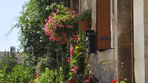 Un edificio con fachada de piedra, contraventanas de madera y macetas con flores rojas.