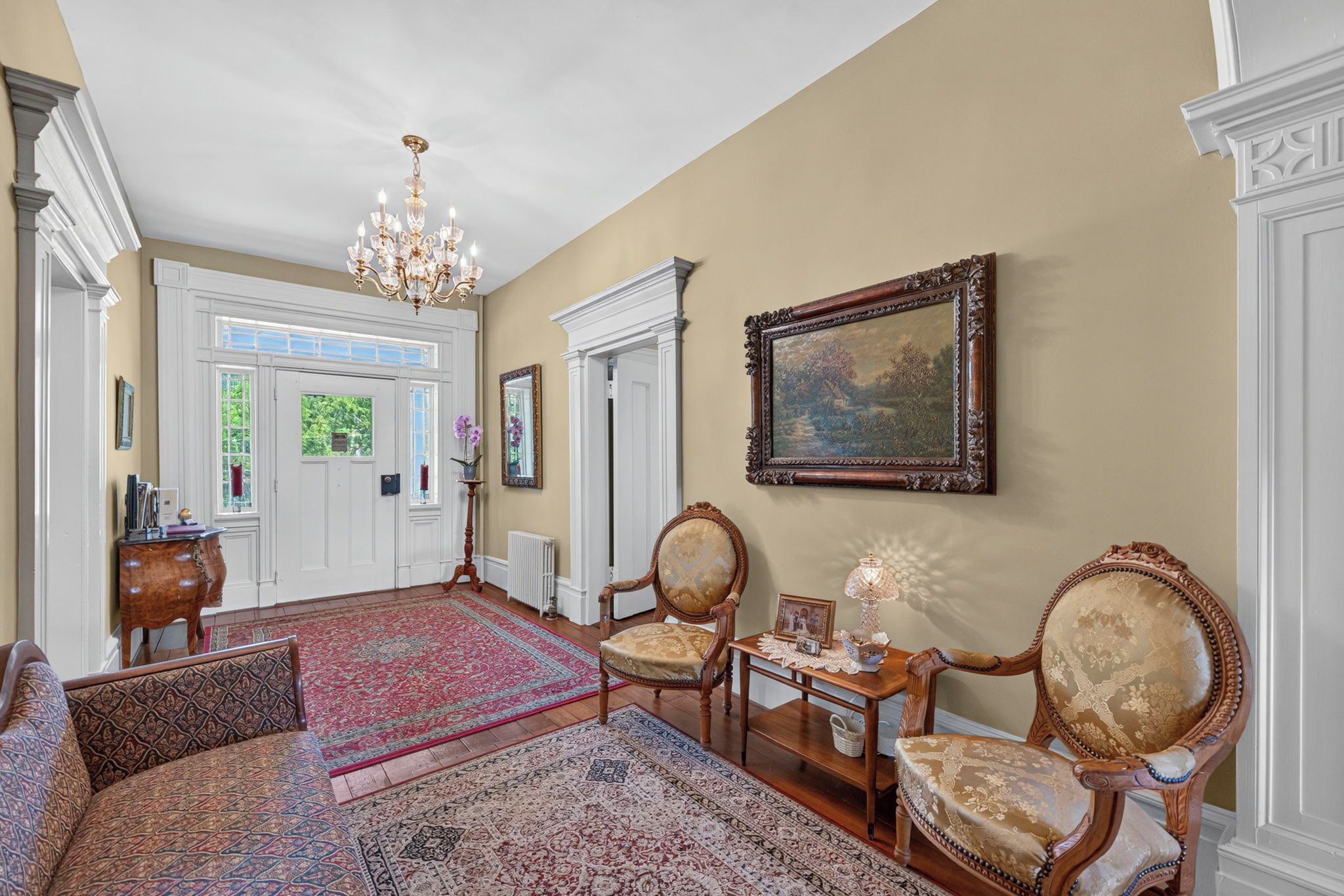 A spacious hallway with a chandelier, ornate chairs, a large rug, and a framed painting on the wall.