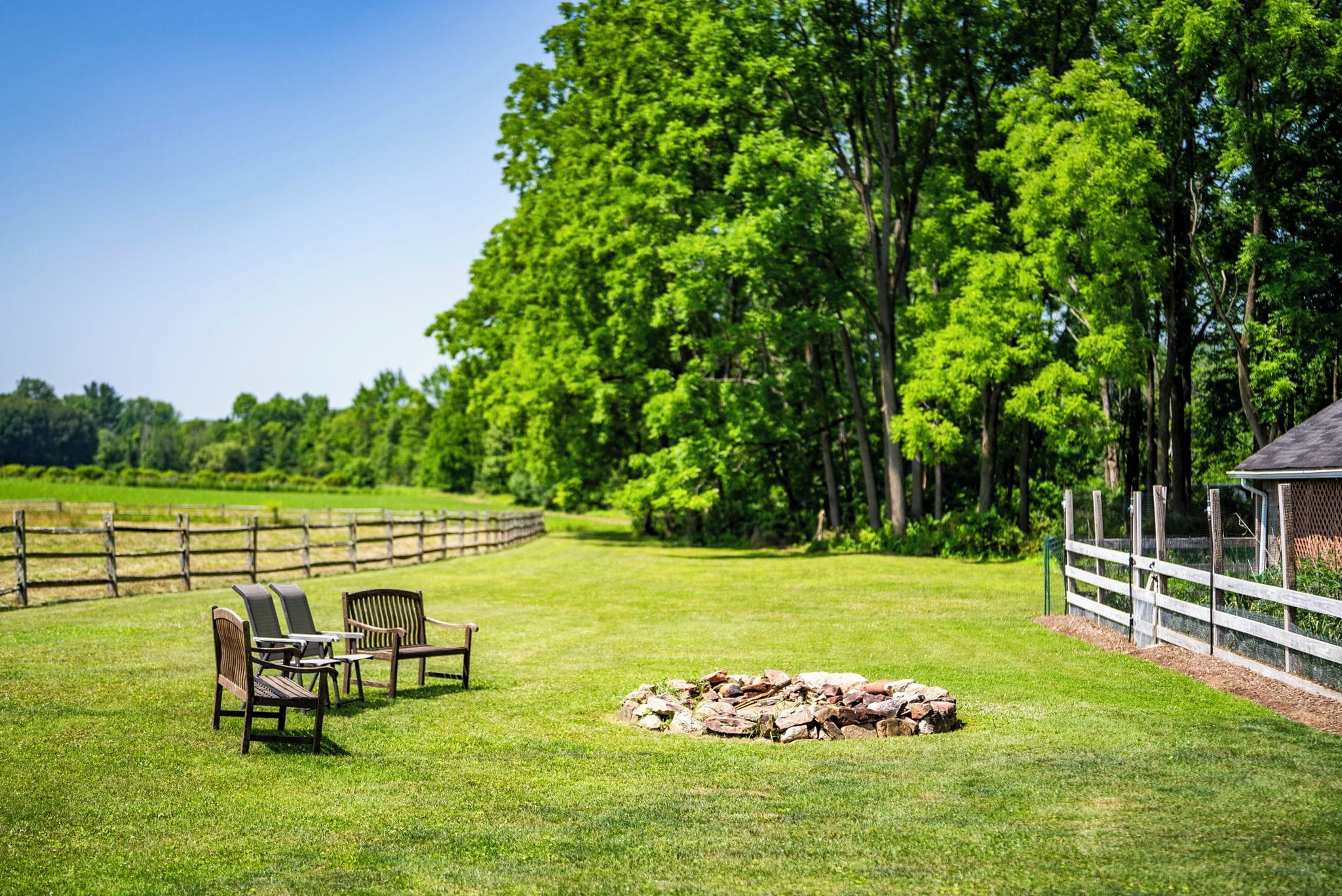 A grassy outdoor area with a wooden fence, a stone fire pit, and two wooden chairs.