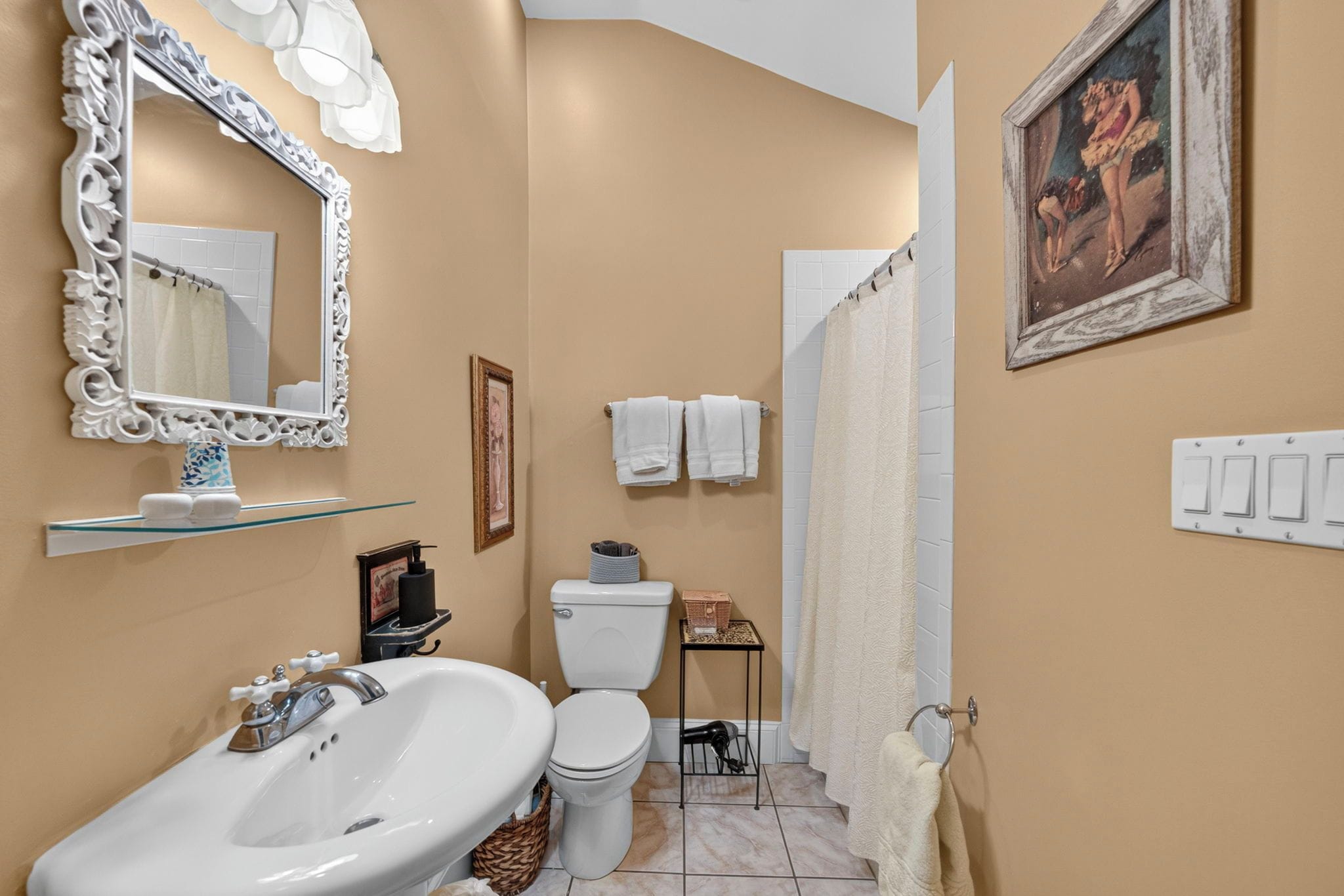 A bathroom with a white sink, toilet, and shower curtain. There is a framed picture on the wall and a shelf with toiletries.