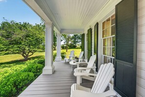 A covered porch with white Adirondack chairs, a small table, and a view of a green landscape. - The Neighbour House B&B (Long Valley)