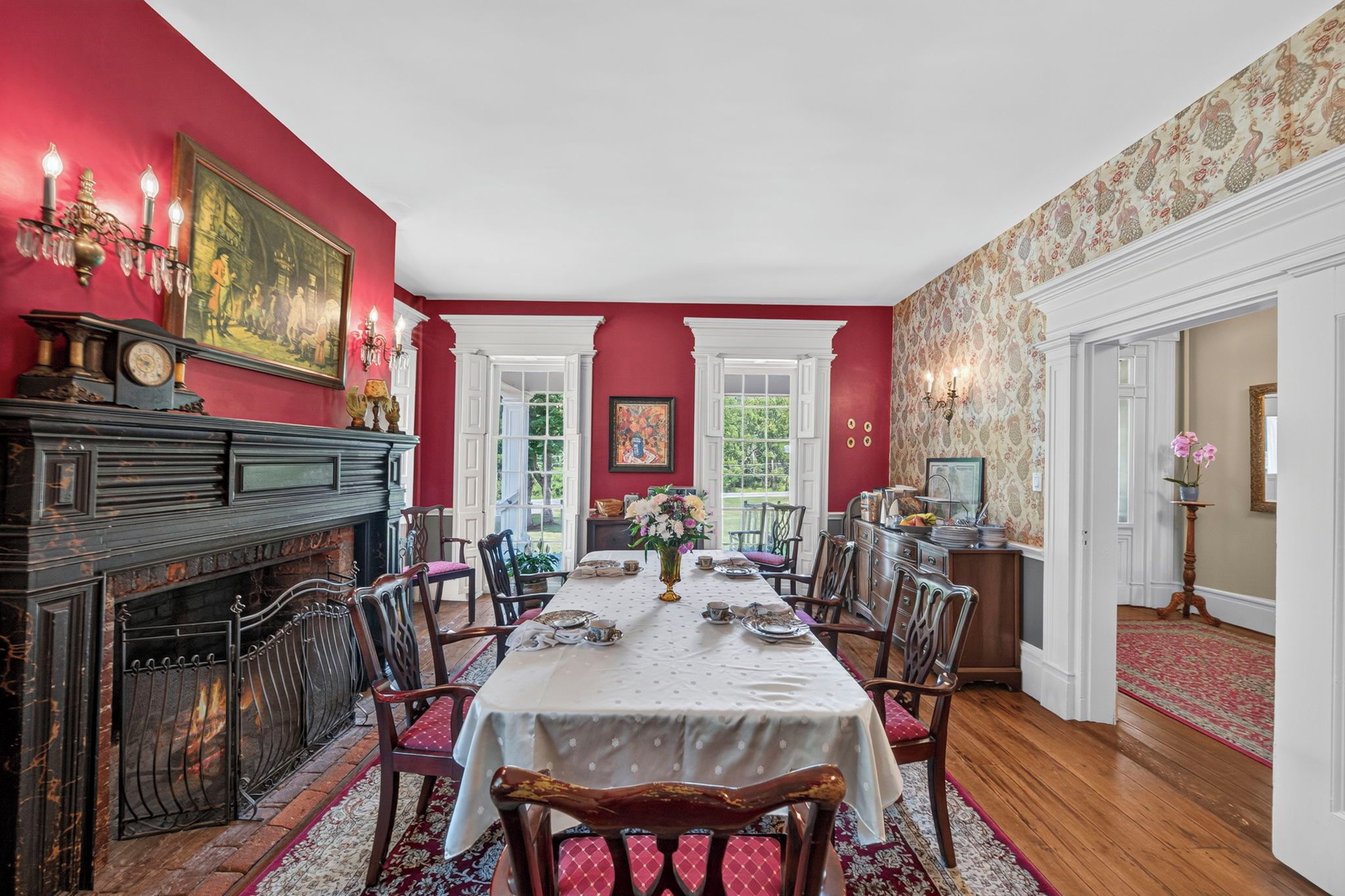 A dining room with a long table set for six, flanked by chairs, a fireplace, and a large painting on the wall.