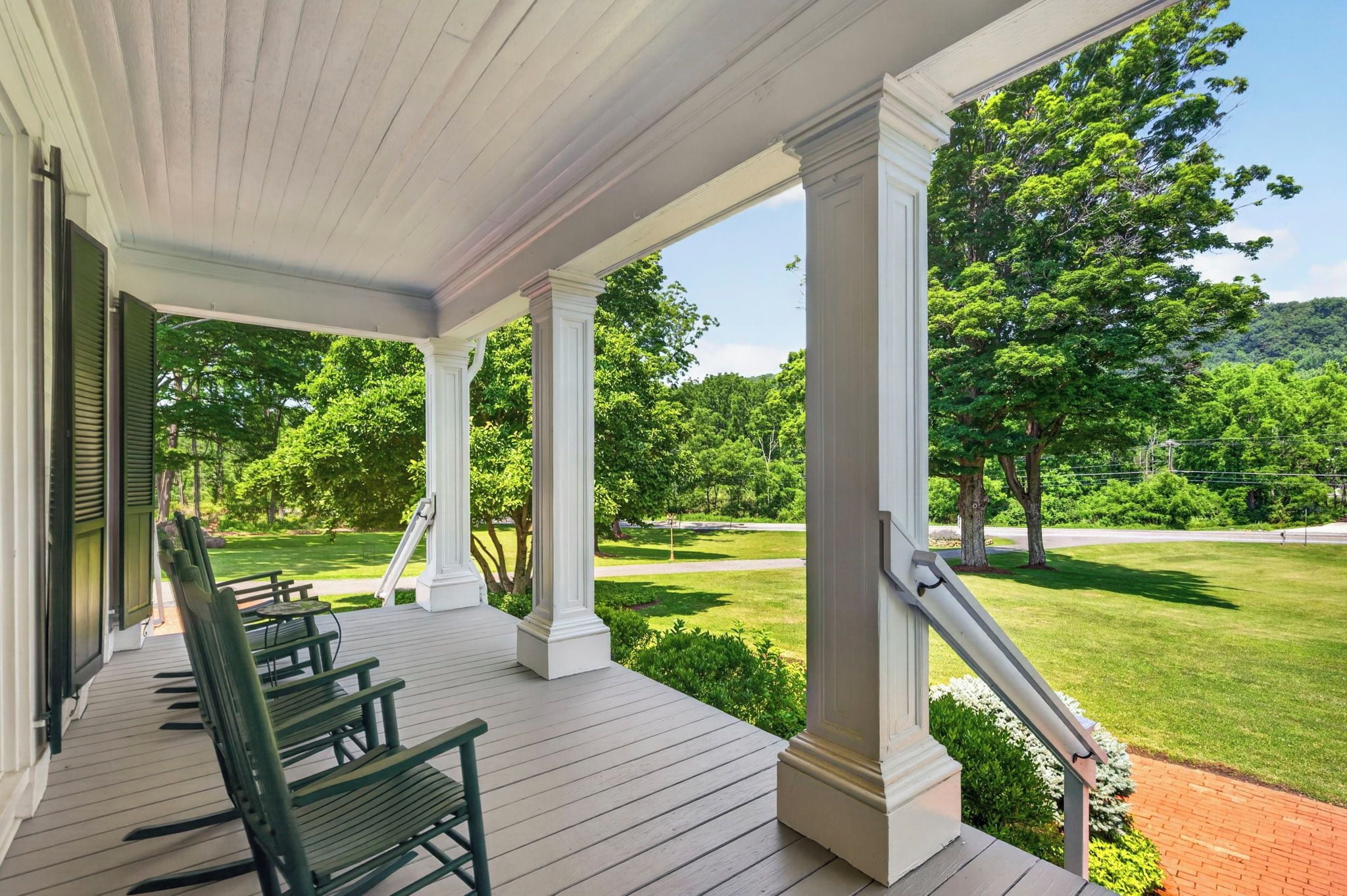 A covered porch with wooden benches, white columns, and a view of a well-maintained lawn and trees.