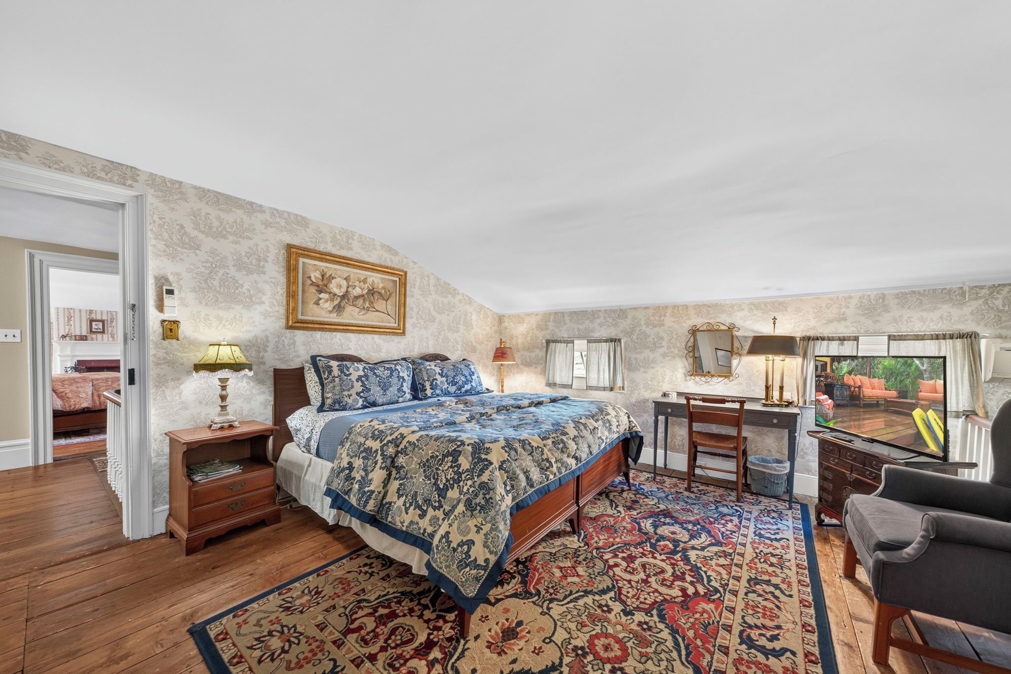 A bedroom with a large bed, a desk, a chair, and a view of the outdoors through a window.