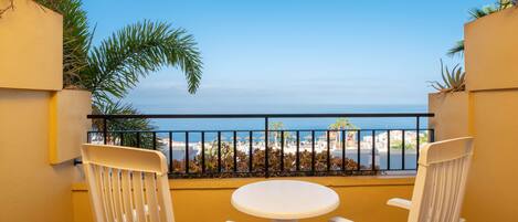Ein Balkon mit zwei weißen Stühlen und einem kleinen runden Tisch, mit Blick auf einen Strand unter blauem Himmel.