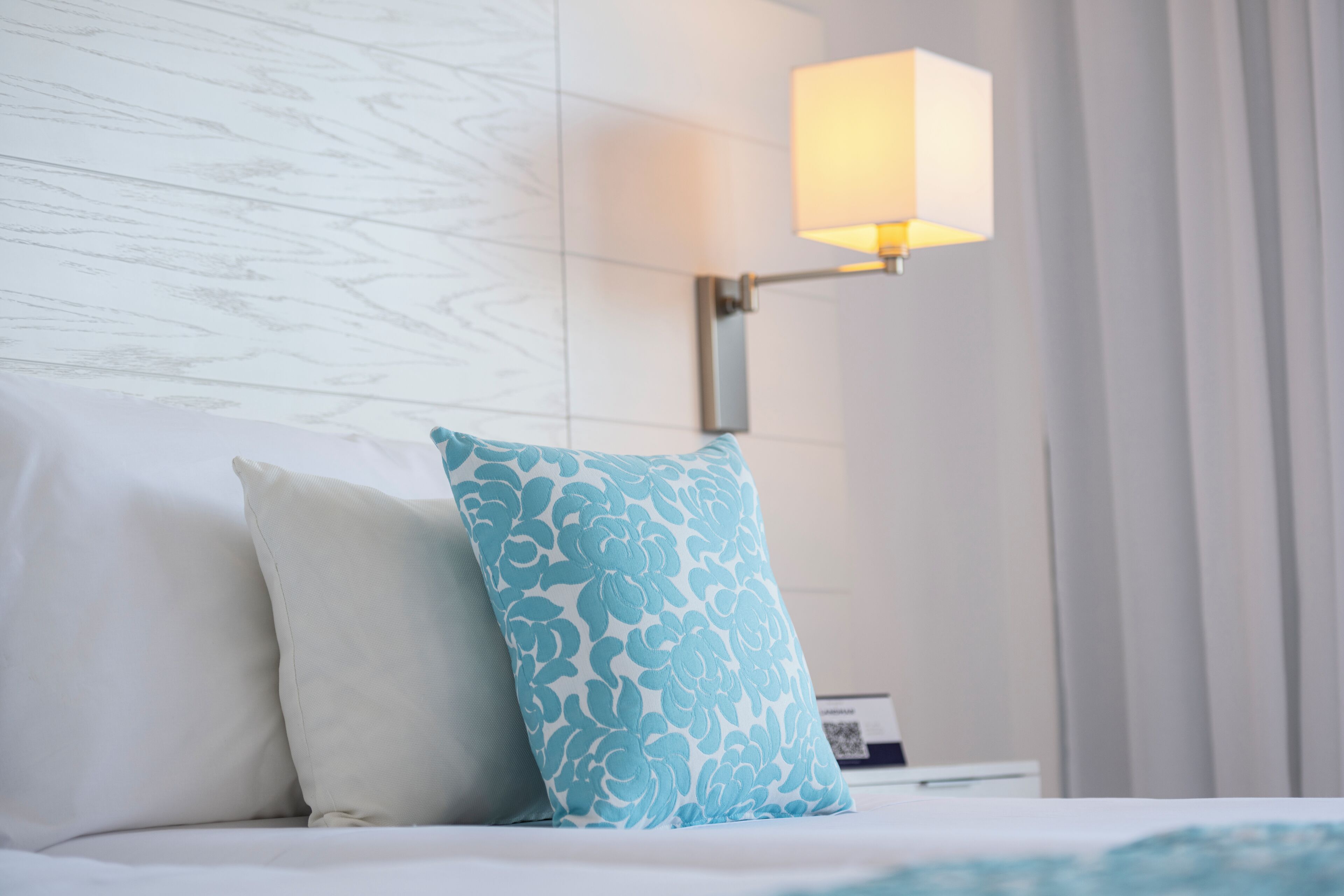 Un lit bien fait, avec deux oreillers : un uni et l’autre à motifs floraux, ainsi qu’une lampe fixée au mur.
