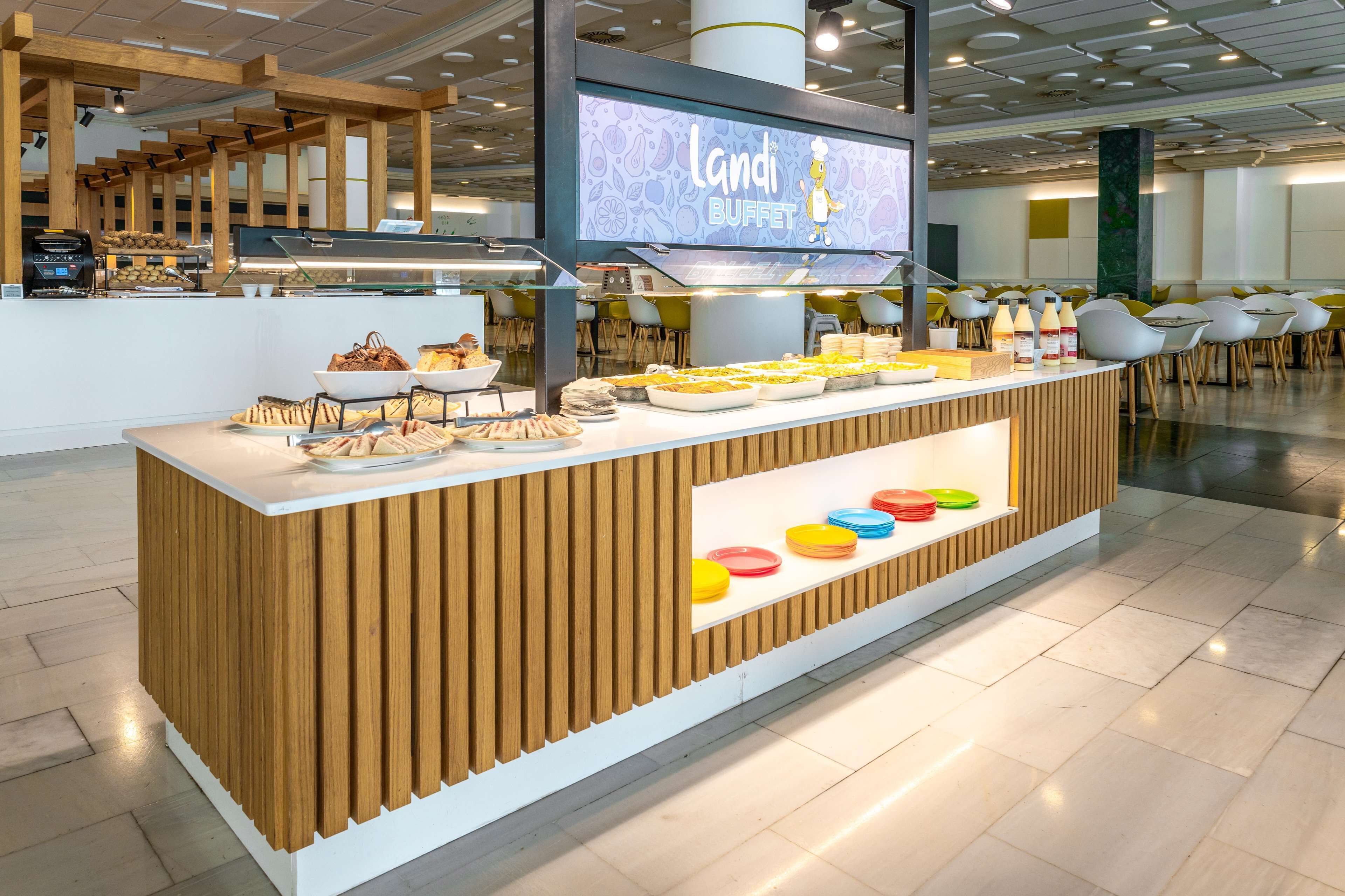 Un espace buffet avec un comptoir en bois, un écran de télévision affichant le mot « LANDI BUFFET », et divers plats proposés.