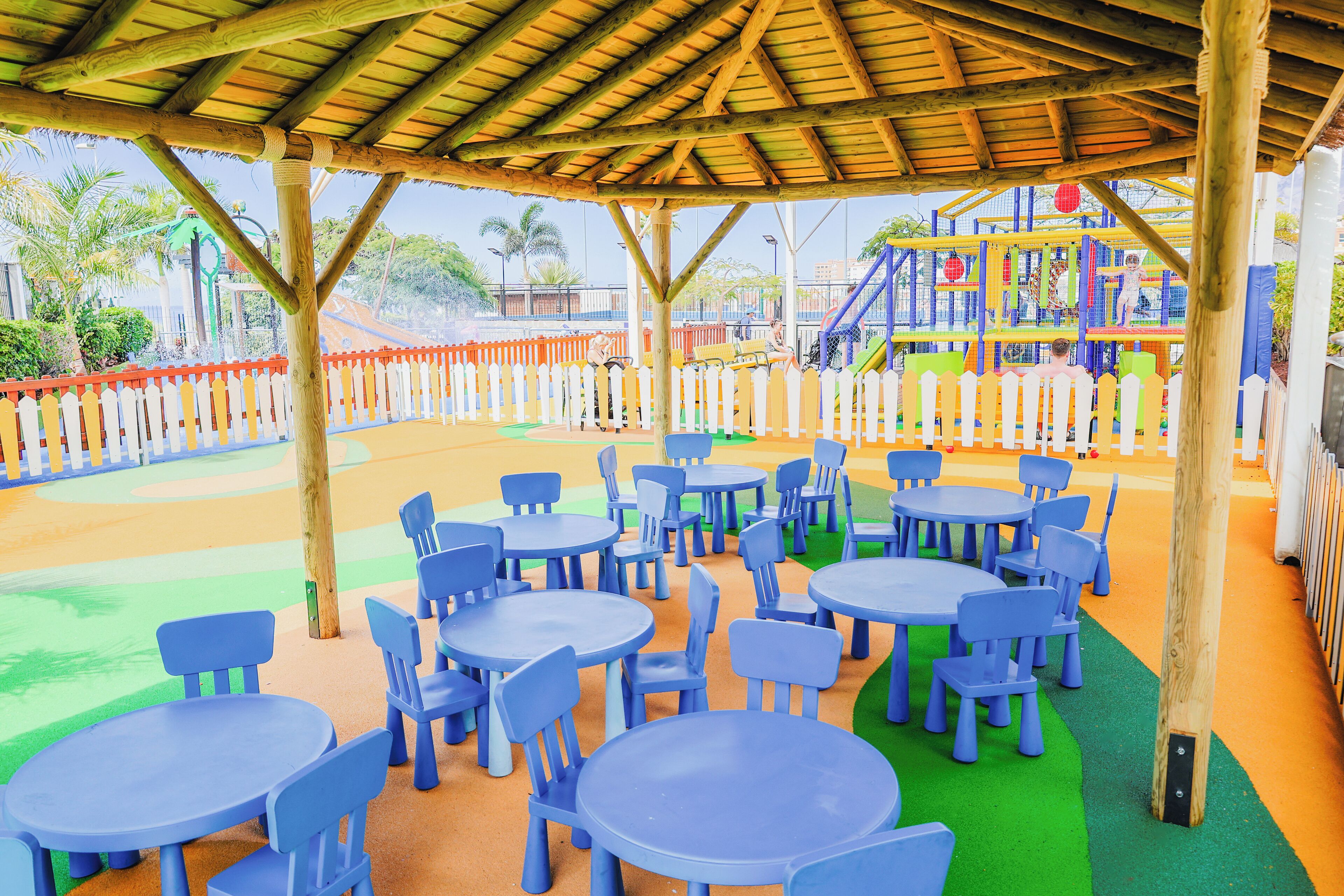 Un espacio al aire libre con mesas y sillas, una estructura de madera sobre él, y un parque infantil vallado al fondo.