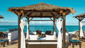 Uma cabana de praia com telhado de palha, cortinas brancas e área de estar acolchoada com vista para o oceano.
