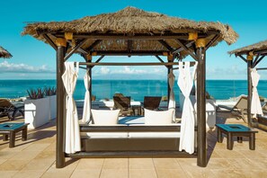 Un cabanon de plage avec un toit de chaume, des rideaux blancs et un coin salon confortable donnant sur l’océan.
