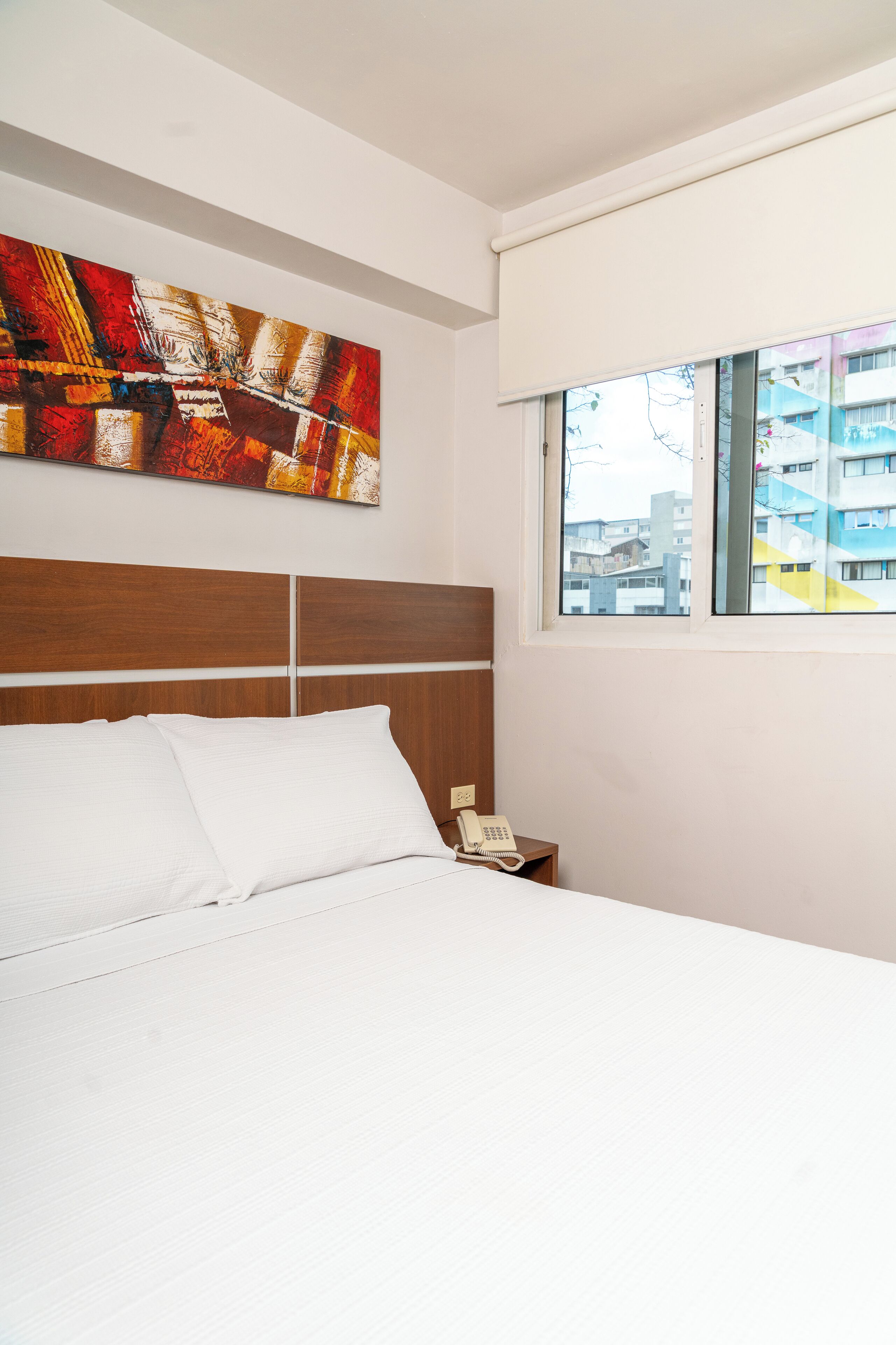 A hotel room with a large bed, a wooden headboard, a bedside telephone, and a window with a city view.
