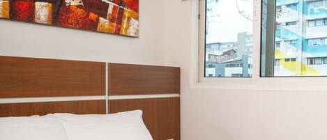 A hotel room with a large bed, a wooden headboard, a bedside telephone, and a window with a city view.