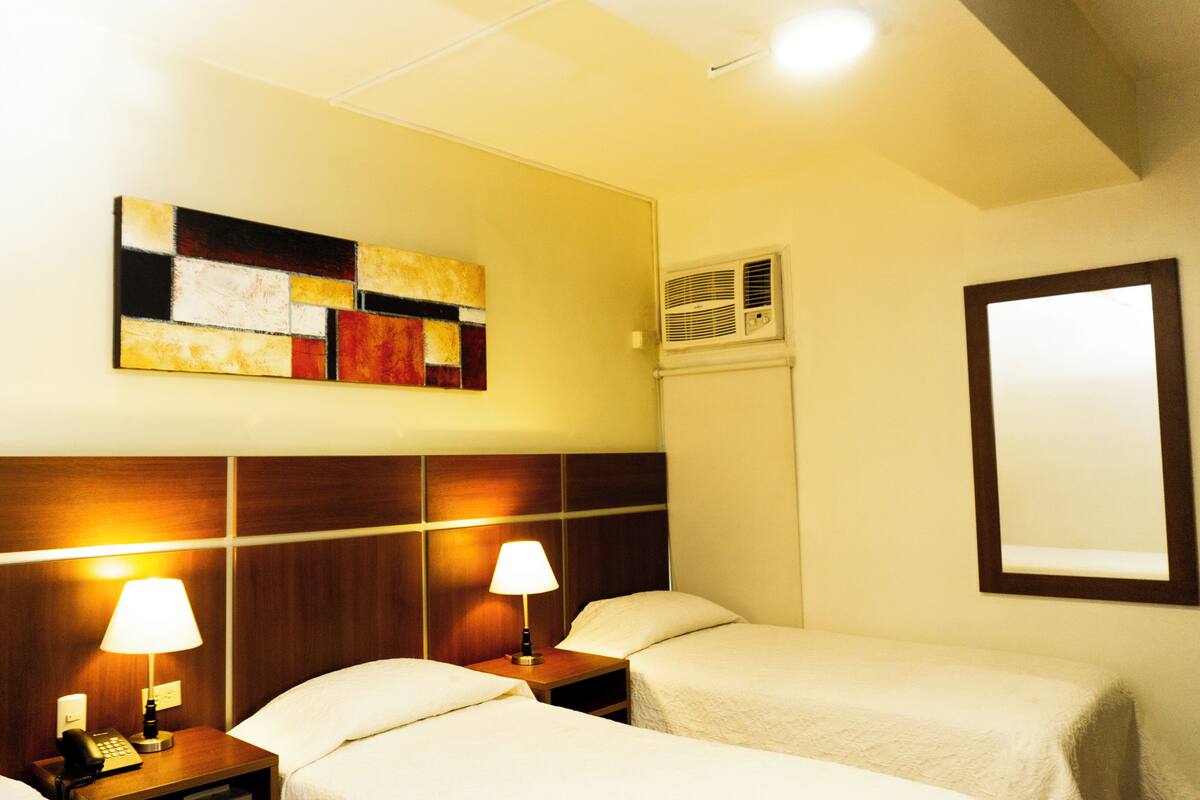 Hotellihuone, jossa on kaksi sänkyä, yöpöytä lampulla, seinään asennettu ilmastointilaite ja suuri peili.