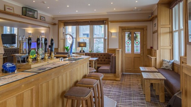 A cozy bar area with wooden bar stools, a counter with beer taps, and a seating area with a sofa and tables. - Lutwidge Arms Limited (Holmrook)
